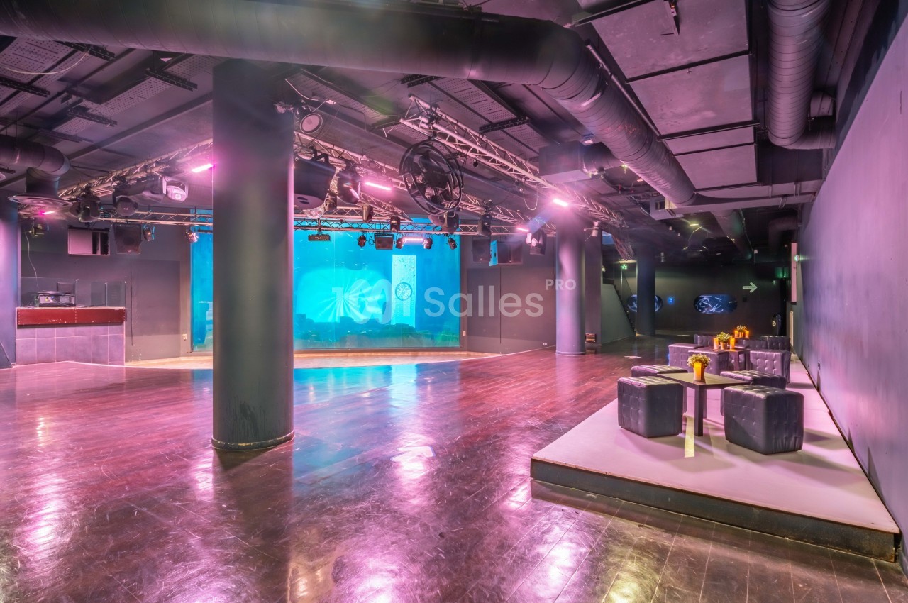 Salle de spectacle vide avec scène éclairée en bleu, sol en parquet et espace lounge avec tables et poufs.