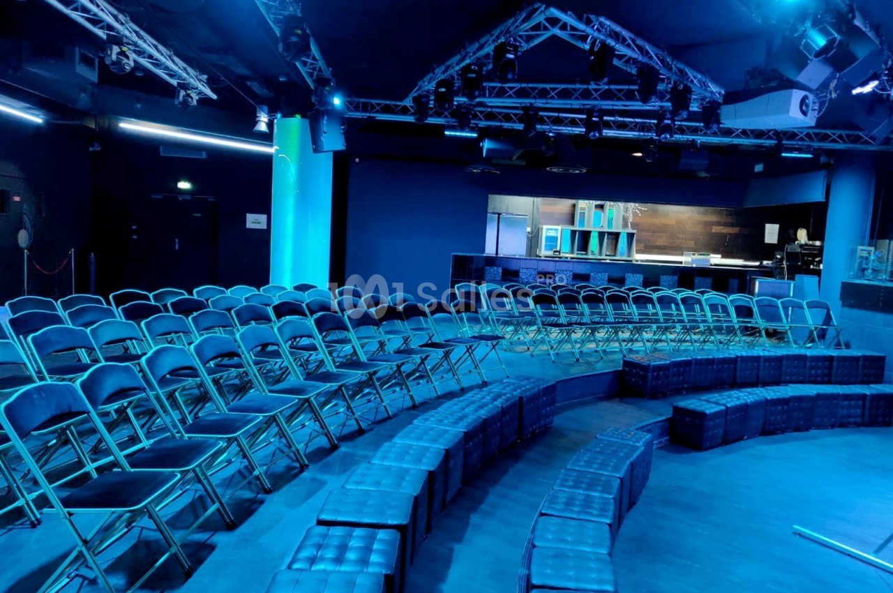 Salle de spectacle avec sièges pliants alignés et banquettes en demi-cercle, éclairée par une lumière bleue.
