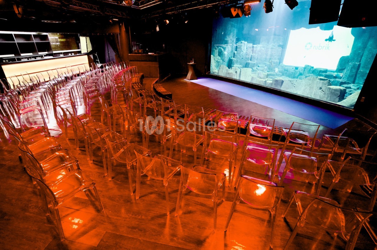 Salle de conférence vide avec des chaises transparentes alignées face à un écran de projection allumé.