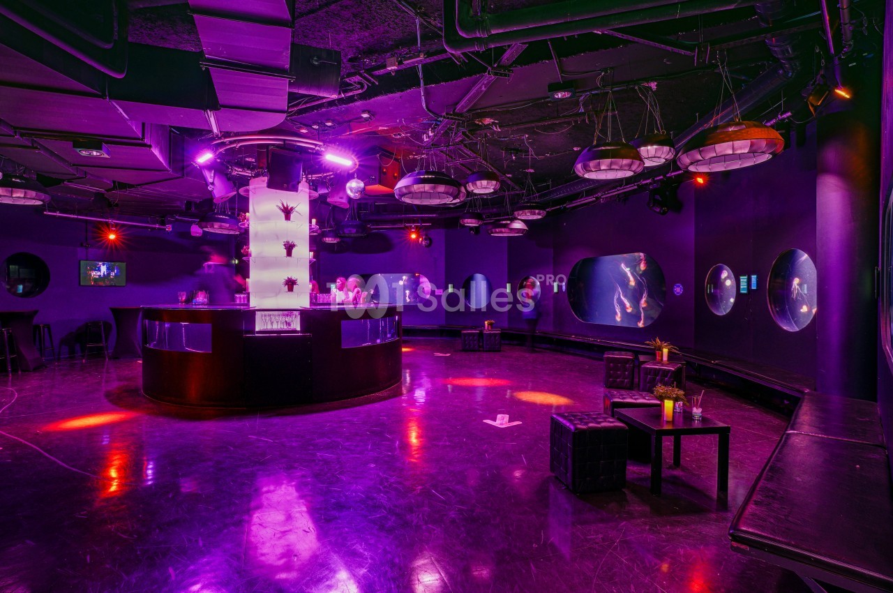 Salle sombre avec éclairage violet, bar central lumineux, tables basses et écrans ronds diffusant des images.