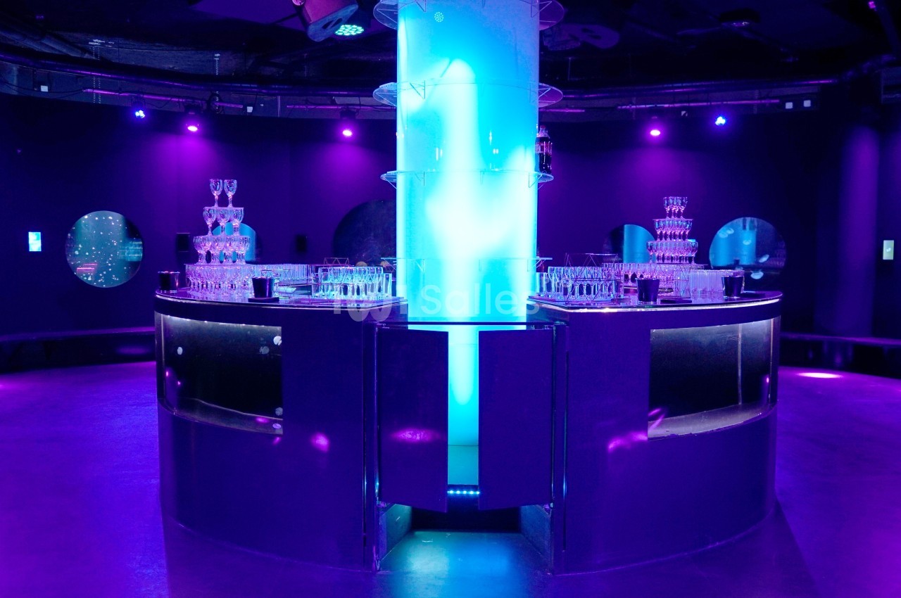 Salle sombre éclairée par des lumières violettes et bleues, avec des tours de verres empilés sur des comptoirs circulaires.