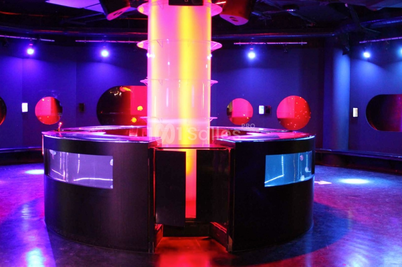 Salle sombre avec éclairage coloré violet et rouge, comptoir central circulaire illuminé et murs avec hublots lumineux.