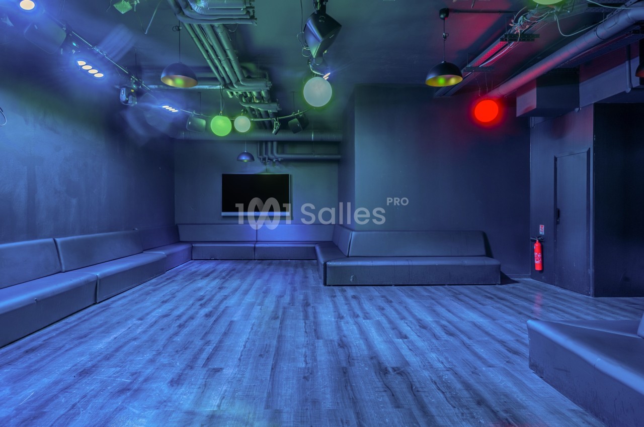 Salle vide avec éclairage tamisé coloré, banquettes noires le long des murs et écran mural.
