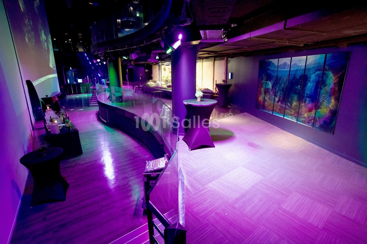 Salle événementielle moderne avec éclairage coloré, tables hautes et décorations, sol en parquet et moquette.