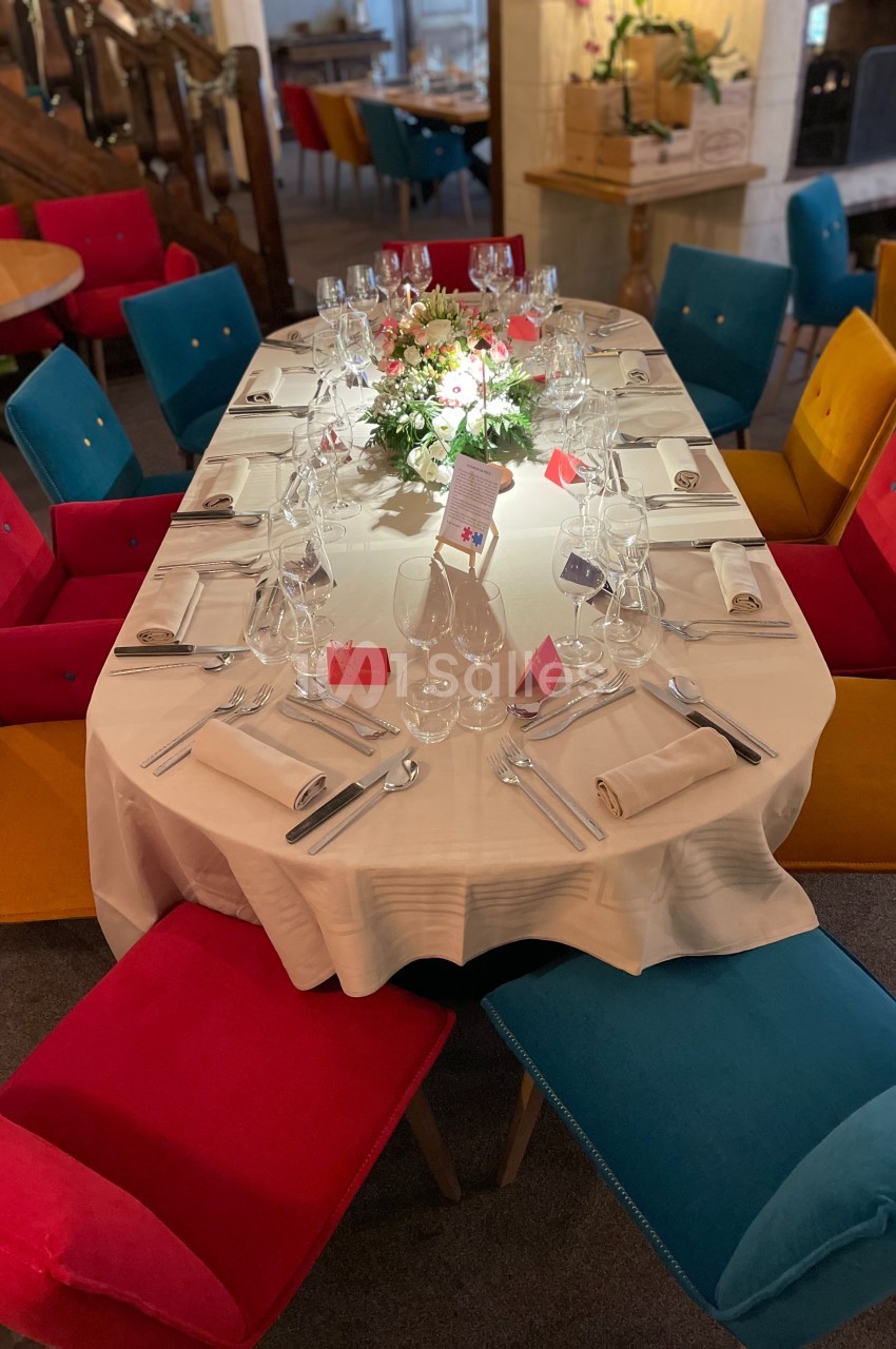 Table ovale dressée avec nappes blanches, verres, couverts et centre de table fleuri, entourée de chaises colorées.
