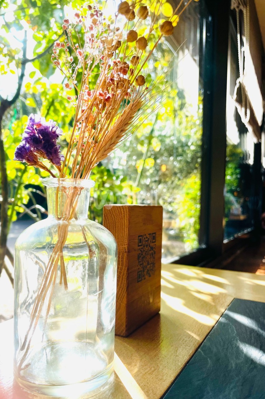Petit vase en verre contenant des fleurs séchées, posé sur une table en bois près d'une fenêtre lumineuse.
