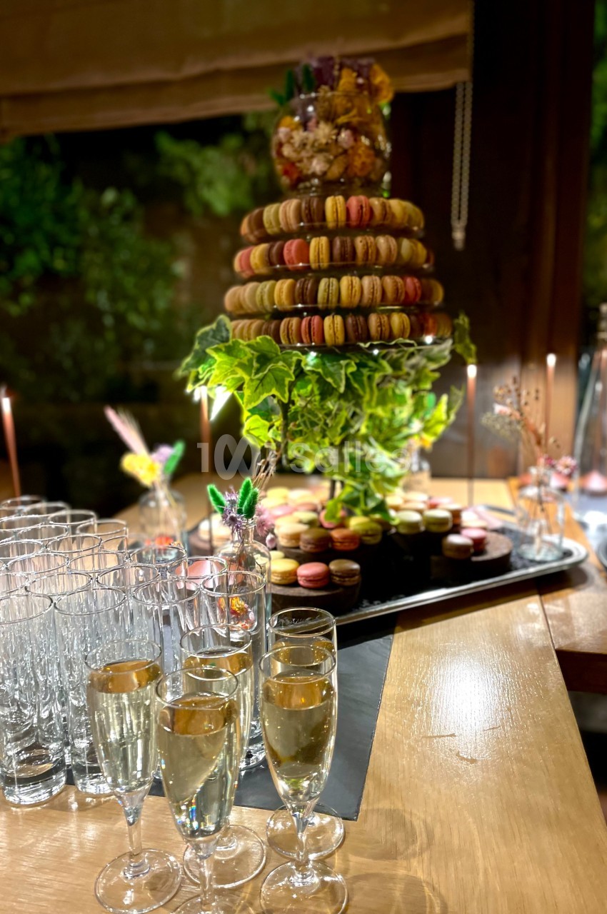 Tour de macarons colorés sur un plateau décoré de verdure, entourée de flûtes de champagne sur une table en bois.