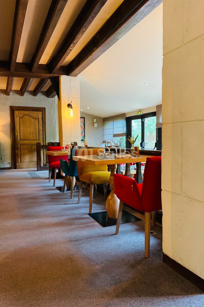 Salle à manger lumineuse avec tables en bois, chaises colorées et poutres apparentes, donnant sur un jardin.