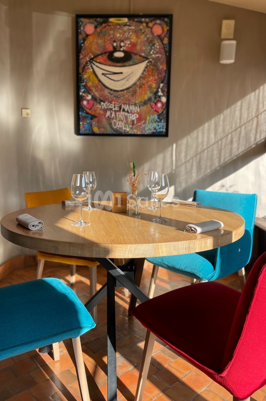 Table ronde dressée avec des verres et serviettes, entourée de chaises colorées, dans une salle lumineuse avec un tableau au…