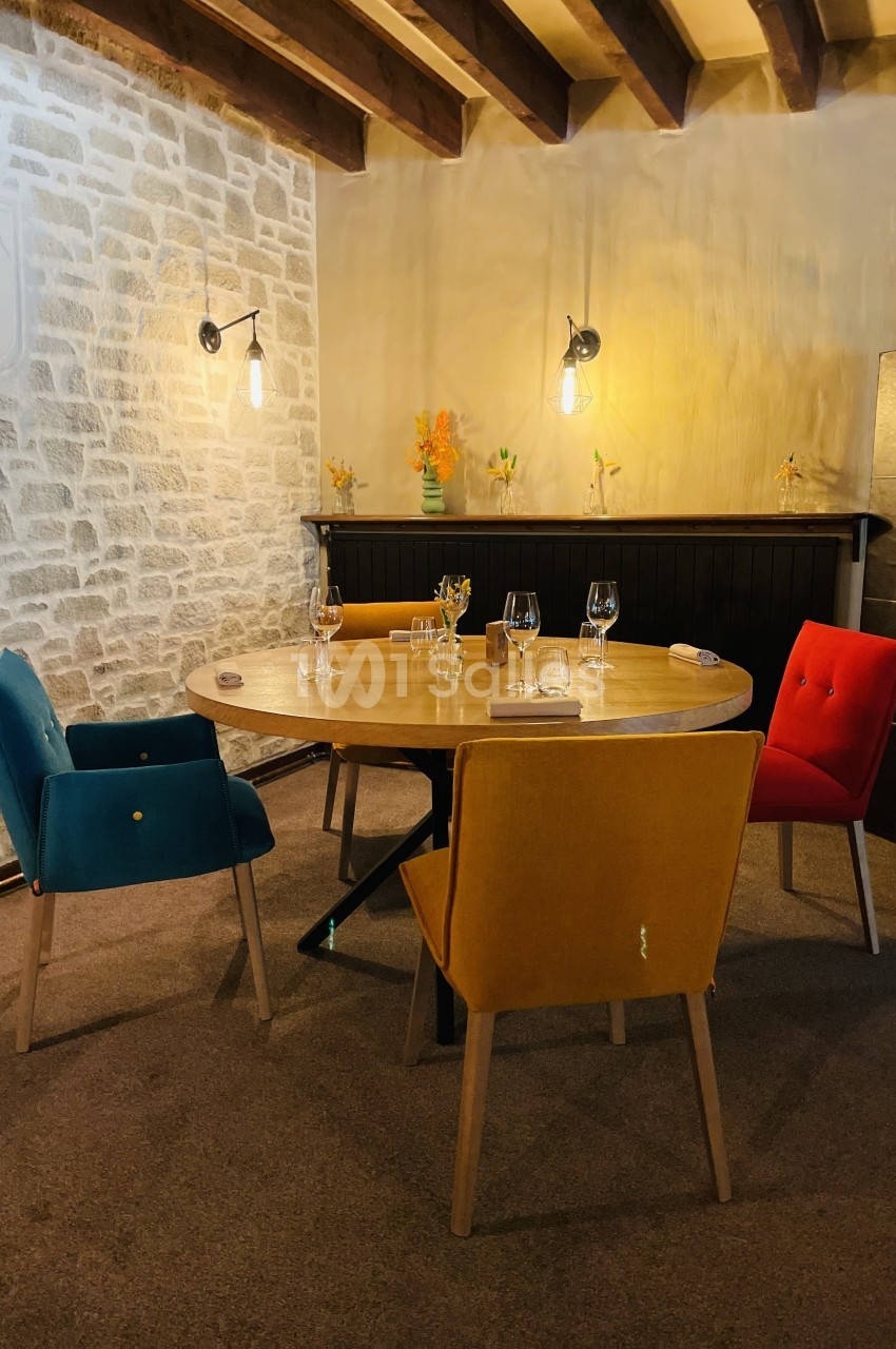 Salle à manger chaleureuse avec table ronde, chaises colorées, murs en pierre et éclairage suspendu.