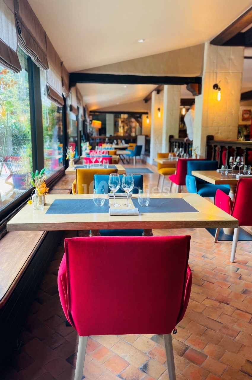 Salle de restaurant lumineuse avec tables dressées, chaises colorées et grandes fenêtres donnant sur un jardin.