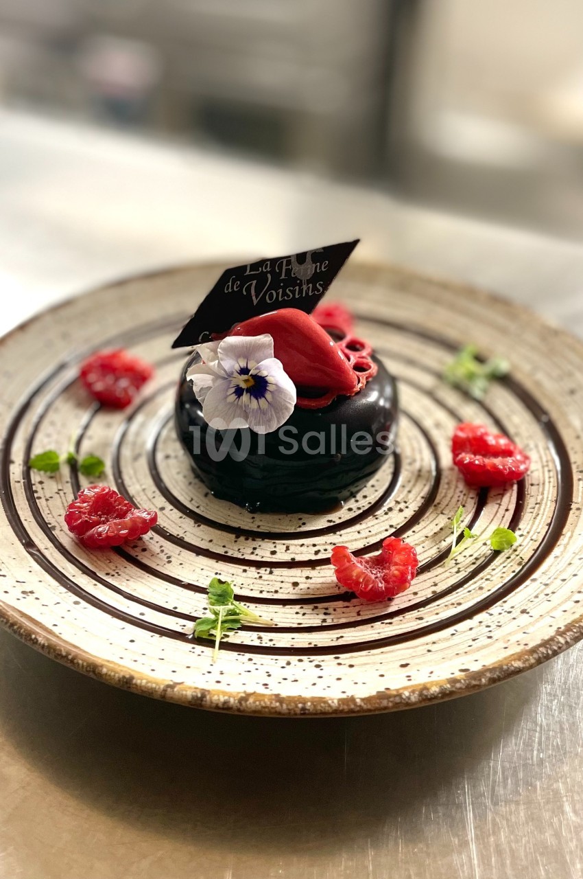 Dessert chocolaté décoré de framboises, fleurs comestibles et feuille en chocolat, présenté sur une assiette élégante.