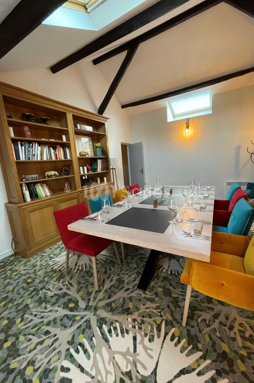 Salle à manger lumineuse avec table dressée, chaises colorées, bibliothèque en bois et poutres apparentes.