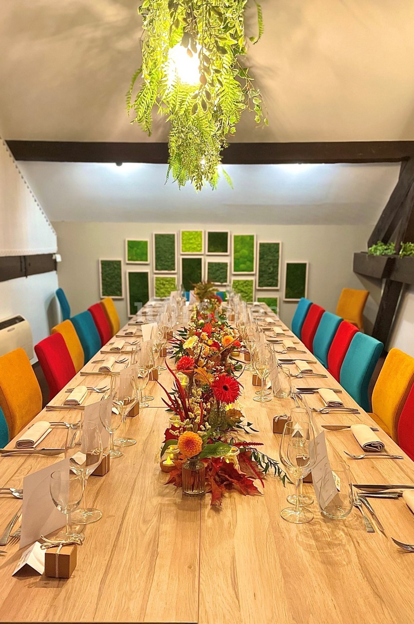 Table en bois dressée pour un repas, décorée de fleurs automnales, entourée de chaises colorées dans une salle lumineuse.