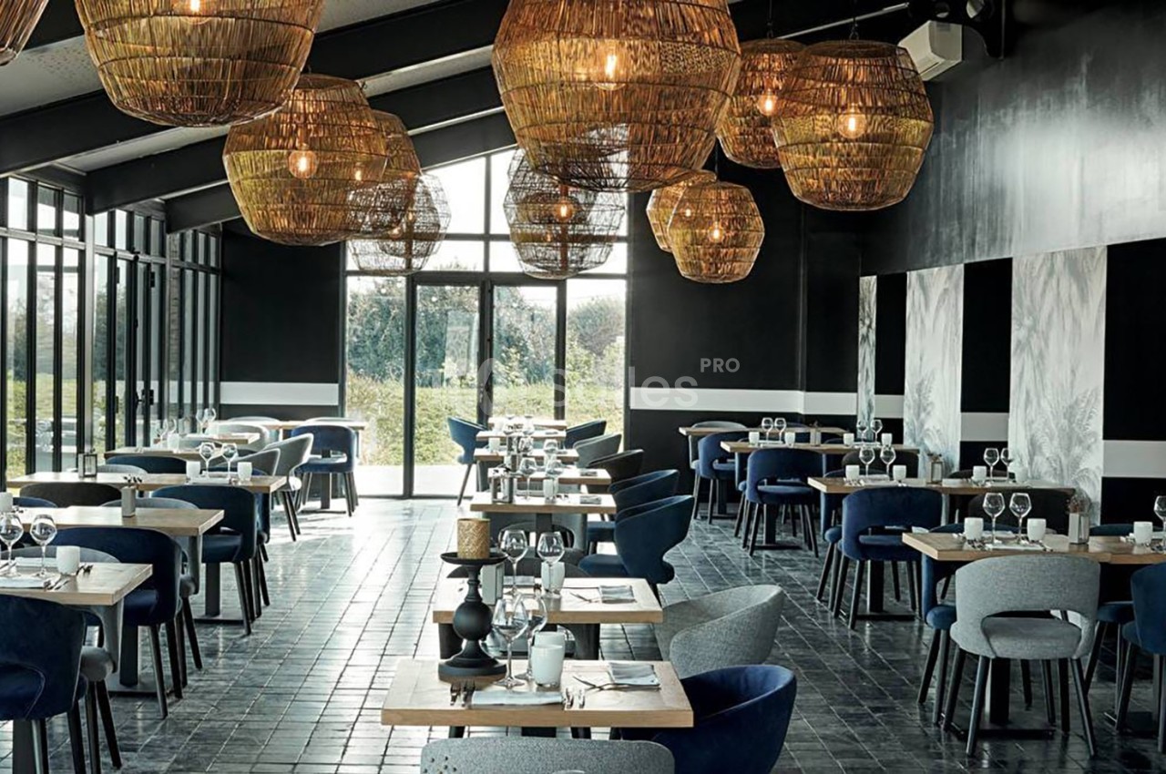 Salle de restaurant moderne avec tables dressées, chaises bleues et grises, grandes baies vitrées et suspensions en osier.
