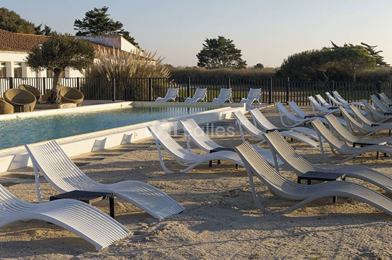 Chaises longues blanches alignées autour d'une piscine extérieure, avec un bâtiment et des arbres en arrière-plan.