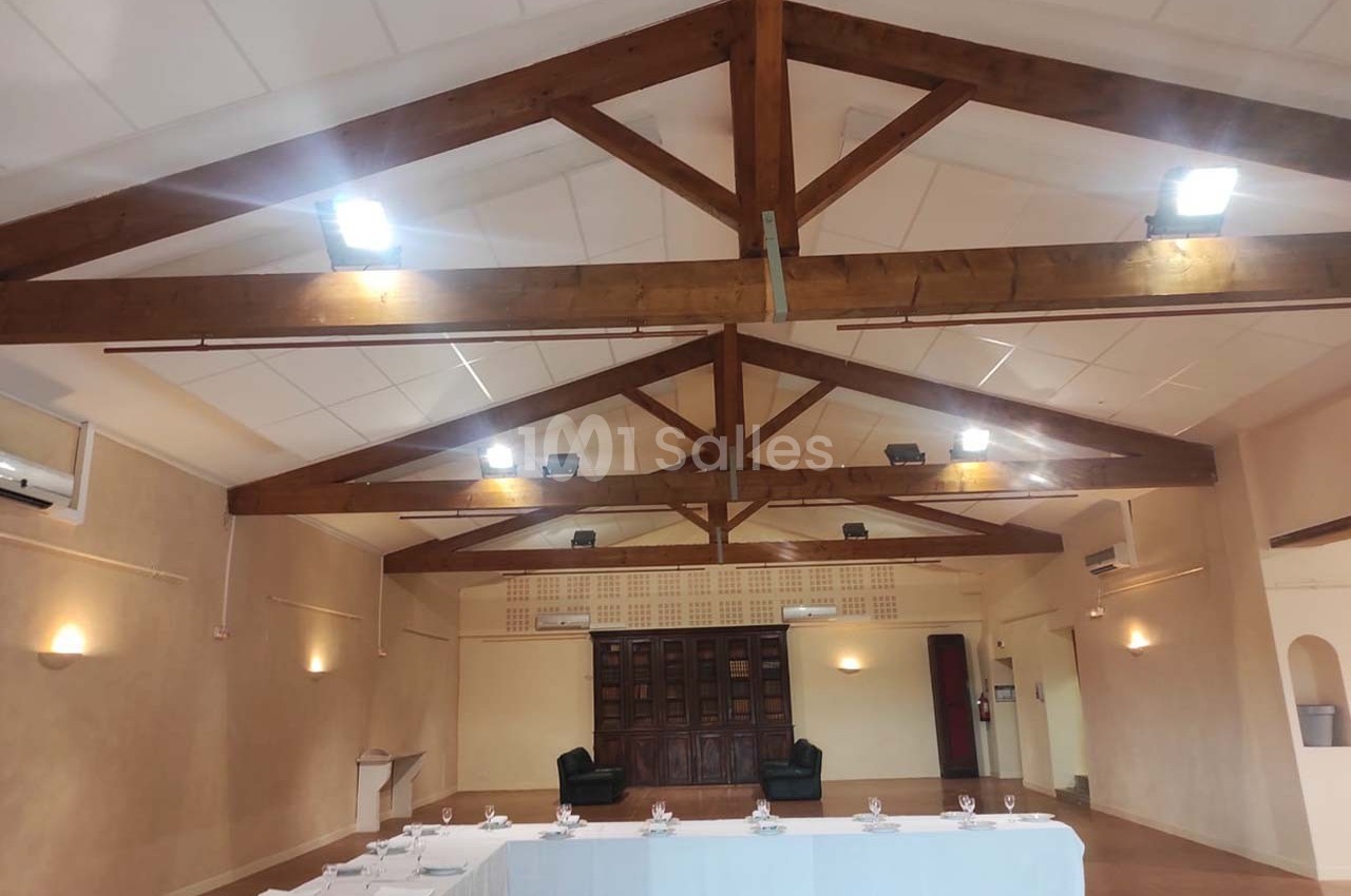 Salle de réception avec poutres apparentes, tables dressées en U et deux fauteuils devant un mur en bois.