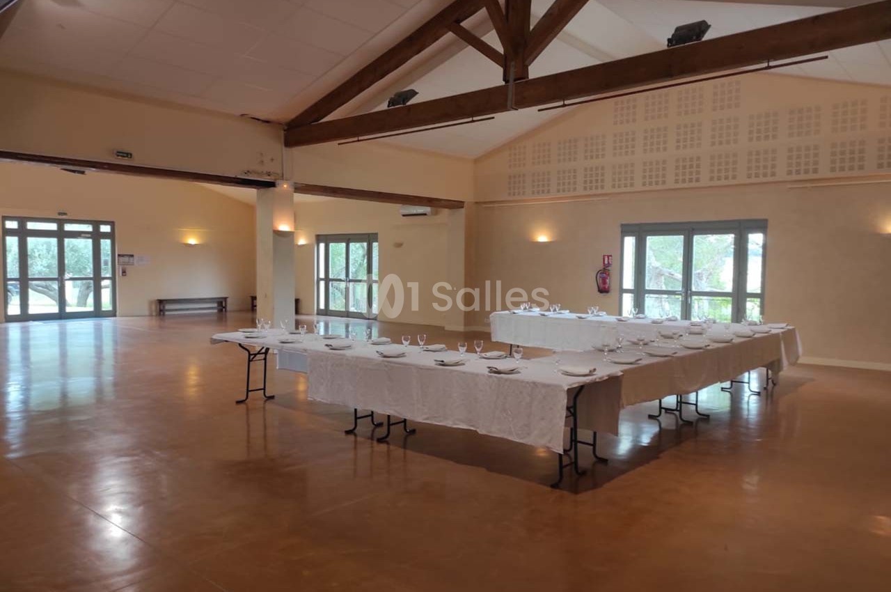 Salle de réception lumineuse avec tables dressées en U, nappes blanches et vaisselle, parquet et poutres apparentes.