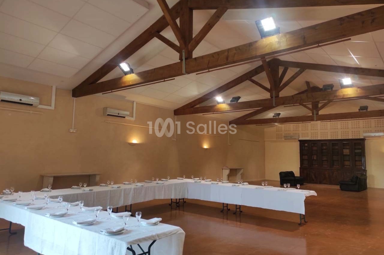 Salle spacieuse avec tables dressées en U, nappes blanches, vaisselle et poutres apparentes au plafond.