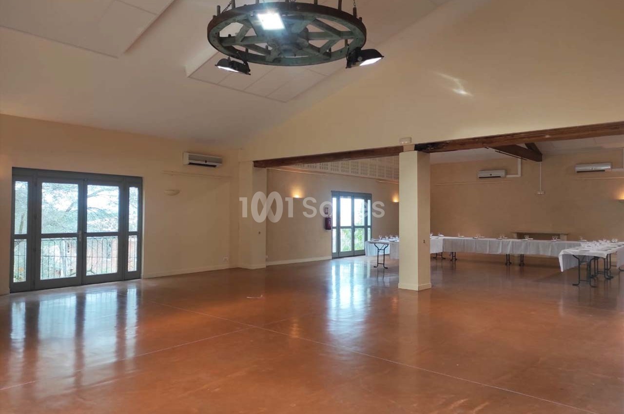 Salle spacieuse avec sol en béton ciré, grandes fenêtres vitrées et tables dressées en arrière-plan.