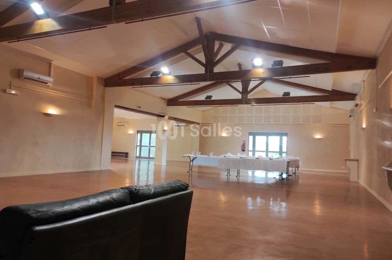 Salle spacieuse avec poutres apparentes, tables dressées au fond et canapé au premier plan.