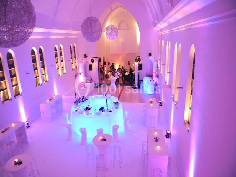 Salle lumineuse avec éclairage violet, tables rondes, chaises transparentes et espace central aménagé pour un événement.
