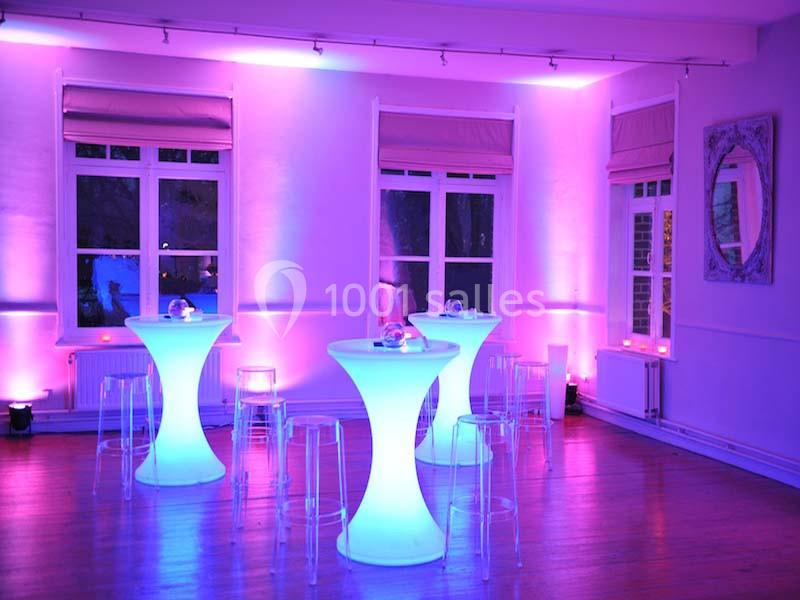 Salle éclairée par des lumières violettes avec des tables hautes lumineuses et des chaises transparentes.