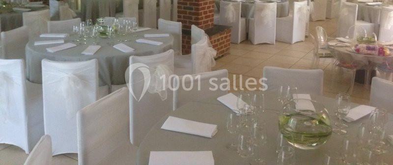Salle décorée pour un événement avec des tables rondes dressées, nappes grises, chaises blanches et centres de table en…