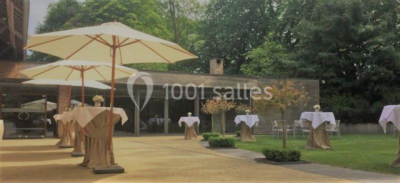 Terrasse aménagée avec des tables hautes couvertes de nappes blanches et des parasols, entourée de verdure.