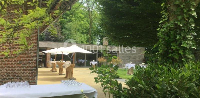 Espace extérieur avec tables dressées sous des parasols, entouré de verdure et d'un bâtiment en bois.
