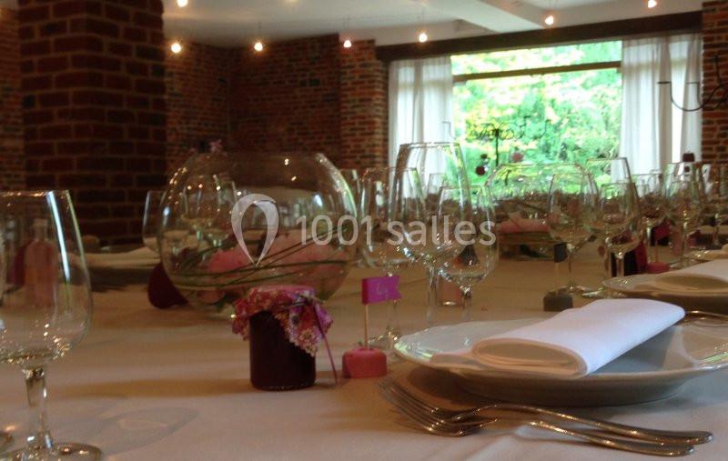 Tables dressées avec nappes blanches, verres, assiettes et décorations florales dans une salle en briques avec vue sur un…