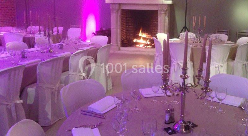 Salle de réception décorée avec des tables dressées, des chaises blanches et une cheminée allumée en arrière-plan.