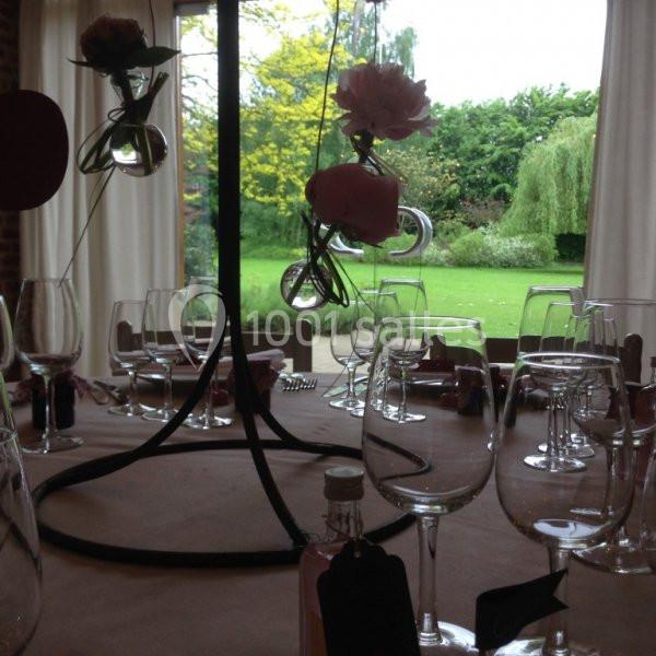 Table décorée avec des verres, des fleurs suspendues et vue sur un jardin verdoyant à travers de grandes fenêtres.