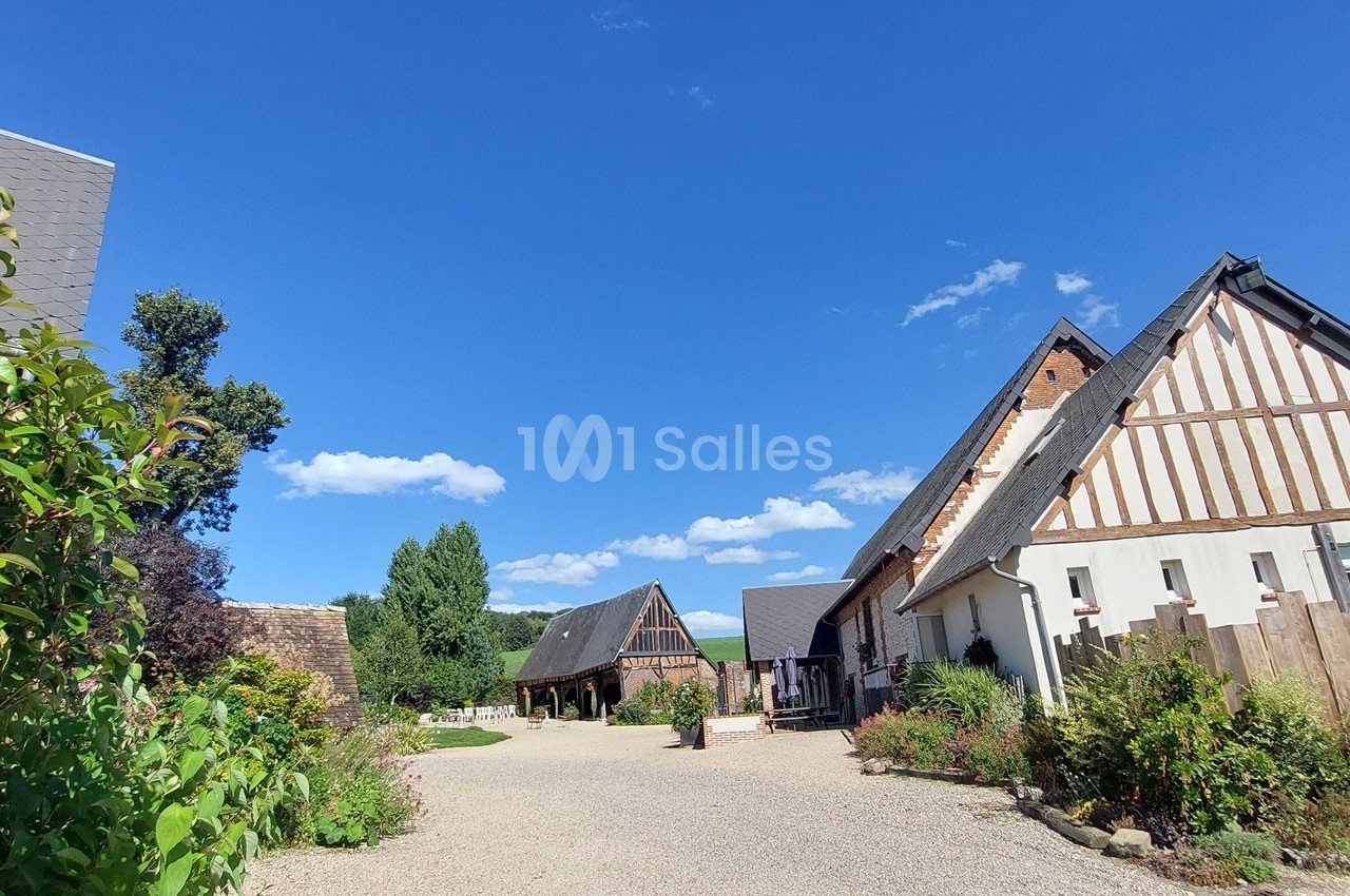 Cour d'une ferme normande avec bâtiments à colombages, jardin fleuri et ciel bleu dégagé en arrière-plan.