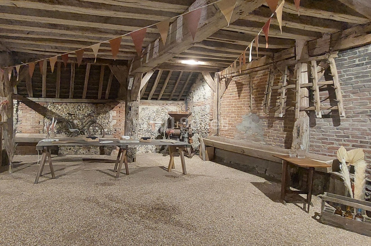 Intérieur d'une grange rustique avec poutres apparentes, guirlandes, outils agricoles et tables en bois.