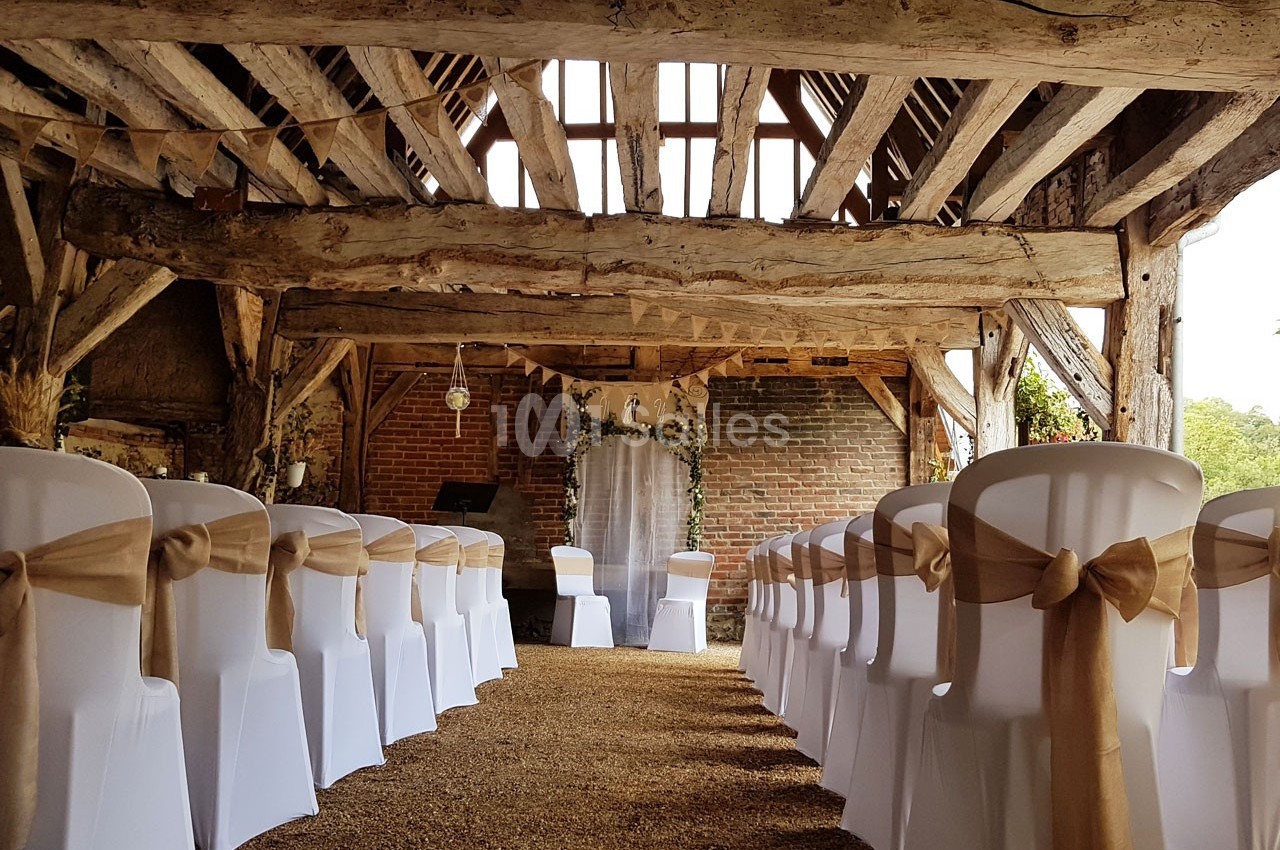 Salle de réception rustique avec des chaises blanches décorées de nœuds en toile de jute, alignées face à une arche fleurie.