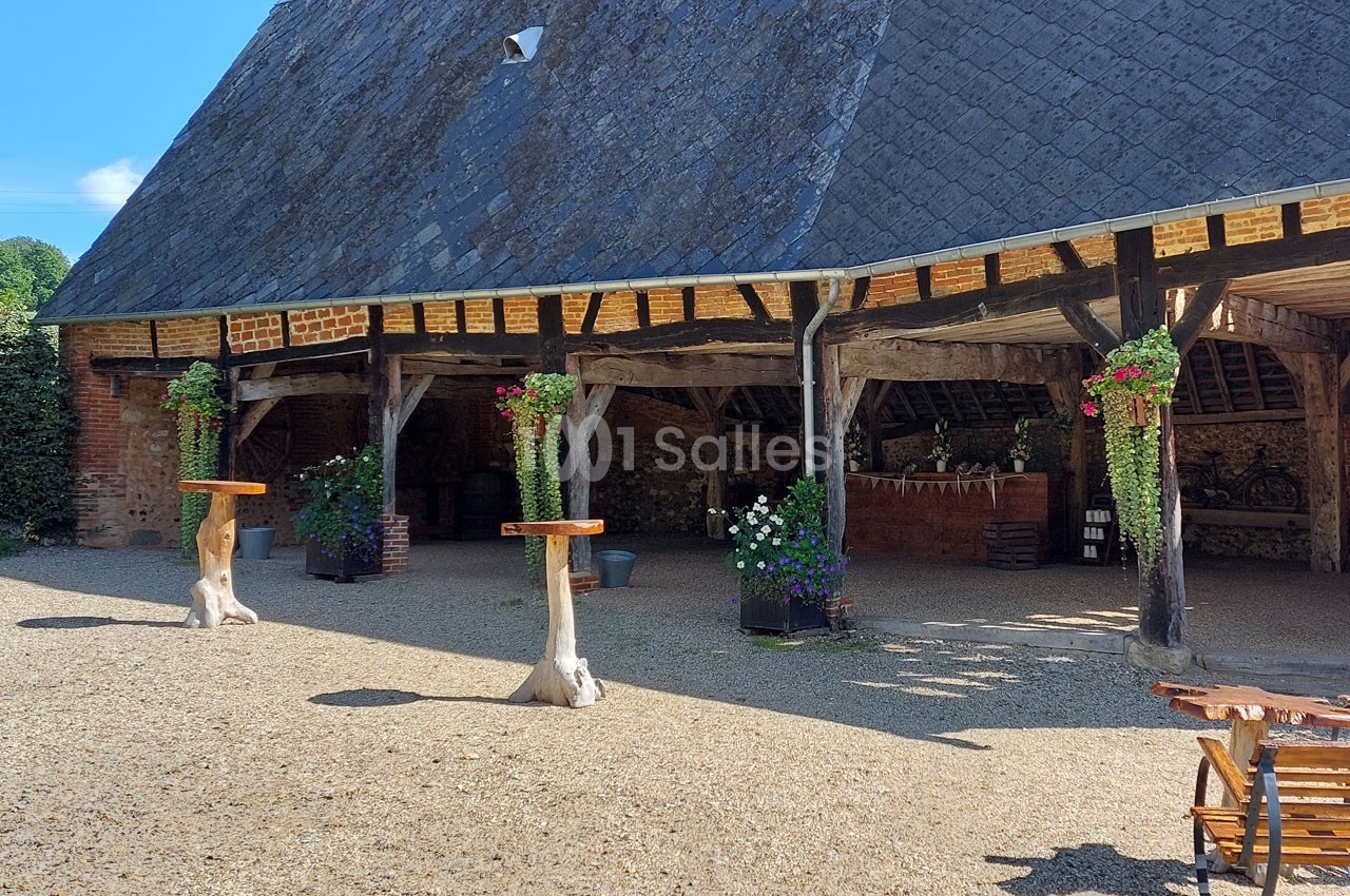 Cour extérieure avec une grange en bois et briques, tables hautes en bois, plantes suspendues et sol en gravier.