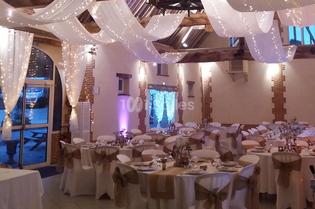 Salle de réception décorée pour un mariage avec tables dressées, chaises ornées de nœuds et guirlandes lumineuses suspendues.