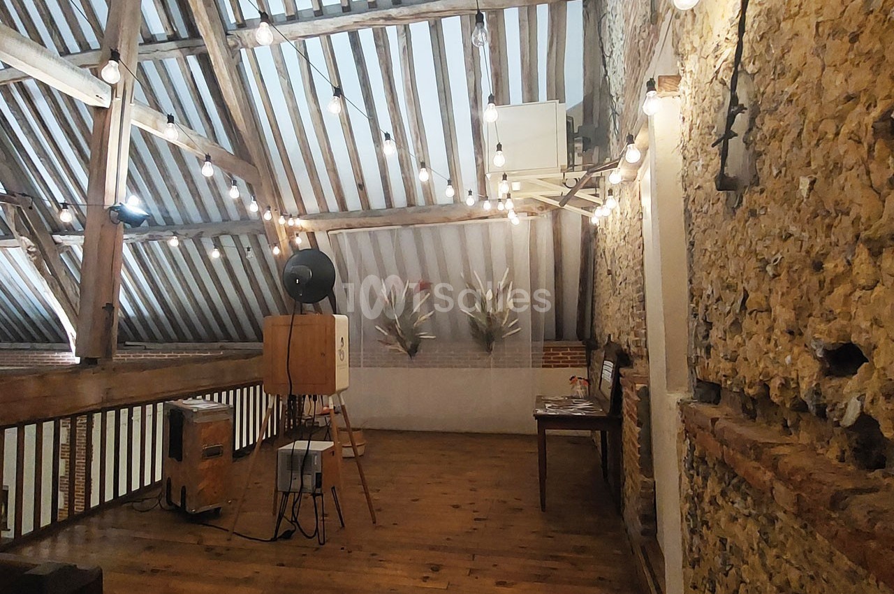 Grenier aménagé avec poutres apparentes, murs en pierre, guirlandes lumineuses et mobilier en bois.