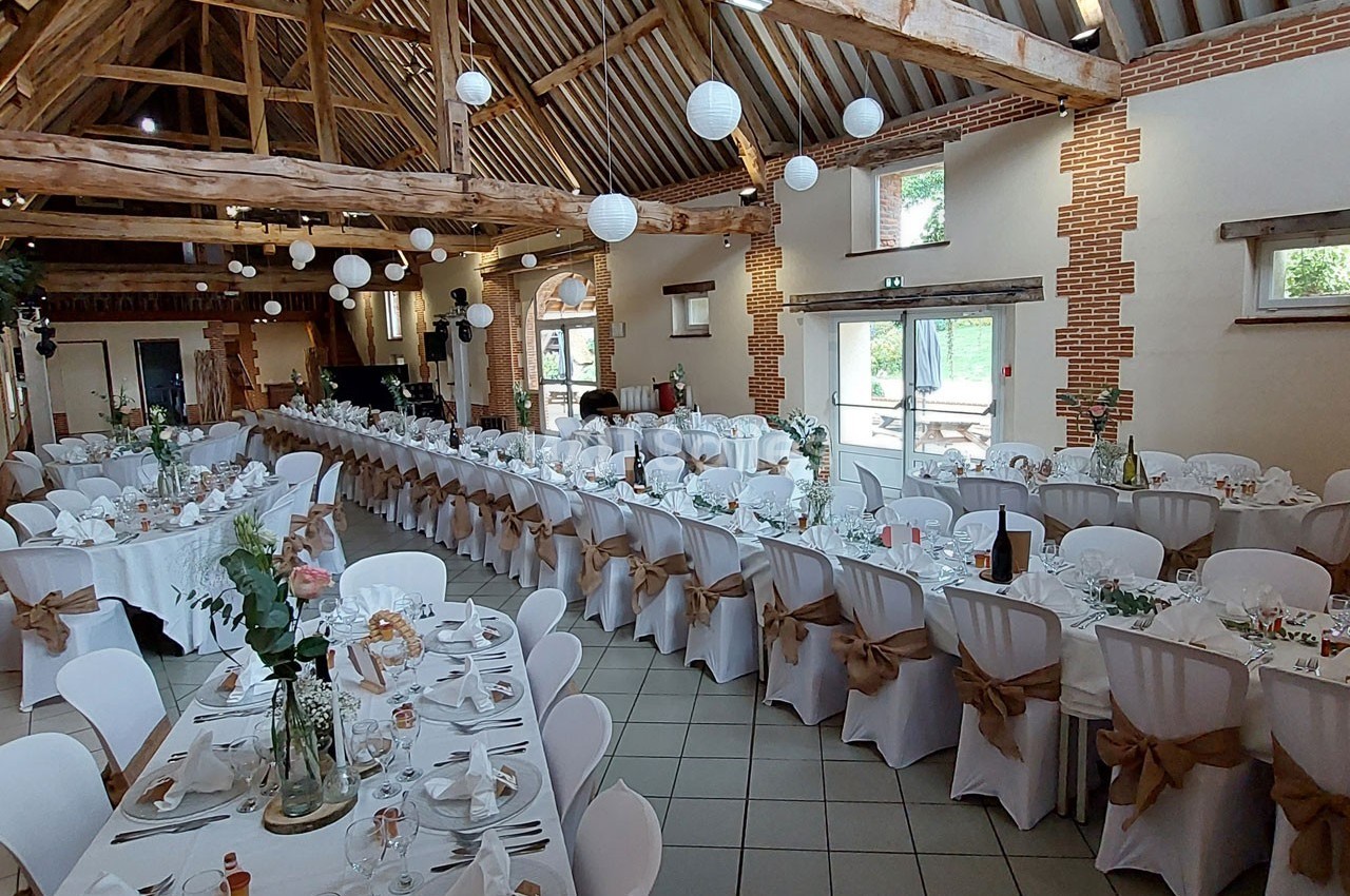 Salle de réception décorée pour un mariage, avec de longues tables dressées, chaises blanches et poutres apparentes.