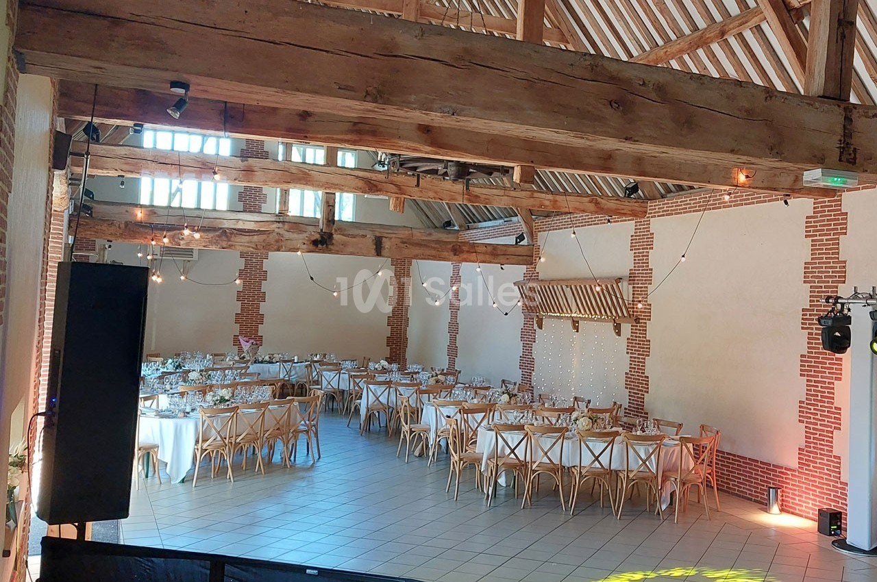 Salle de réception lumineuse avec poutres apparentes, tables dressées et chaises en bois dans un décor rustique.