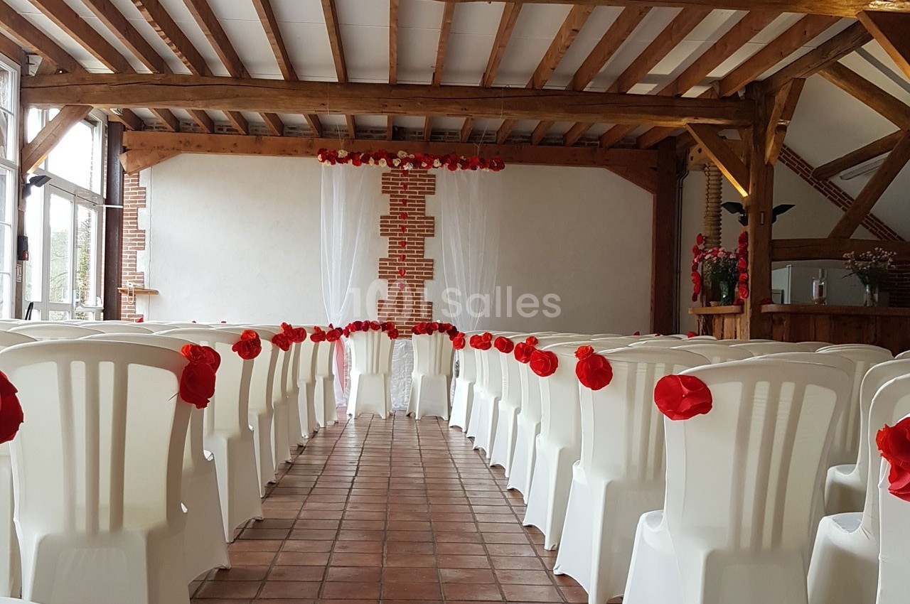 Salle décorée pour une cérémonie, avec des chaises blanches ornées de fleurs rouges alignées face à un mur en briques.