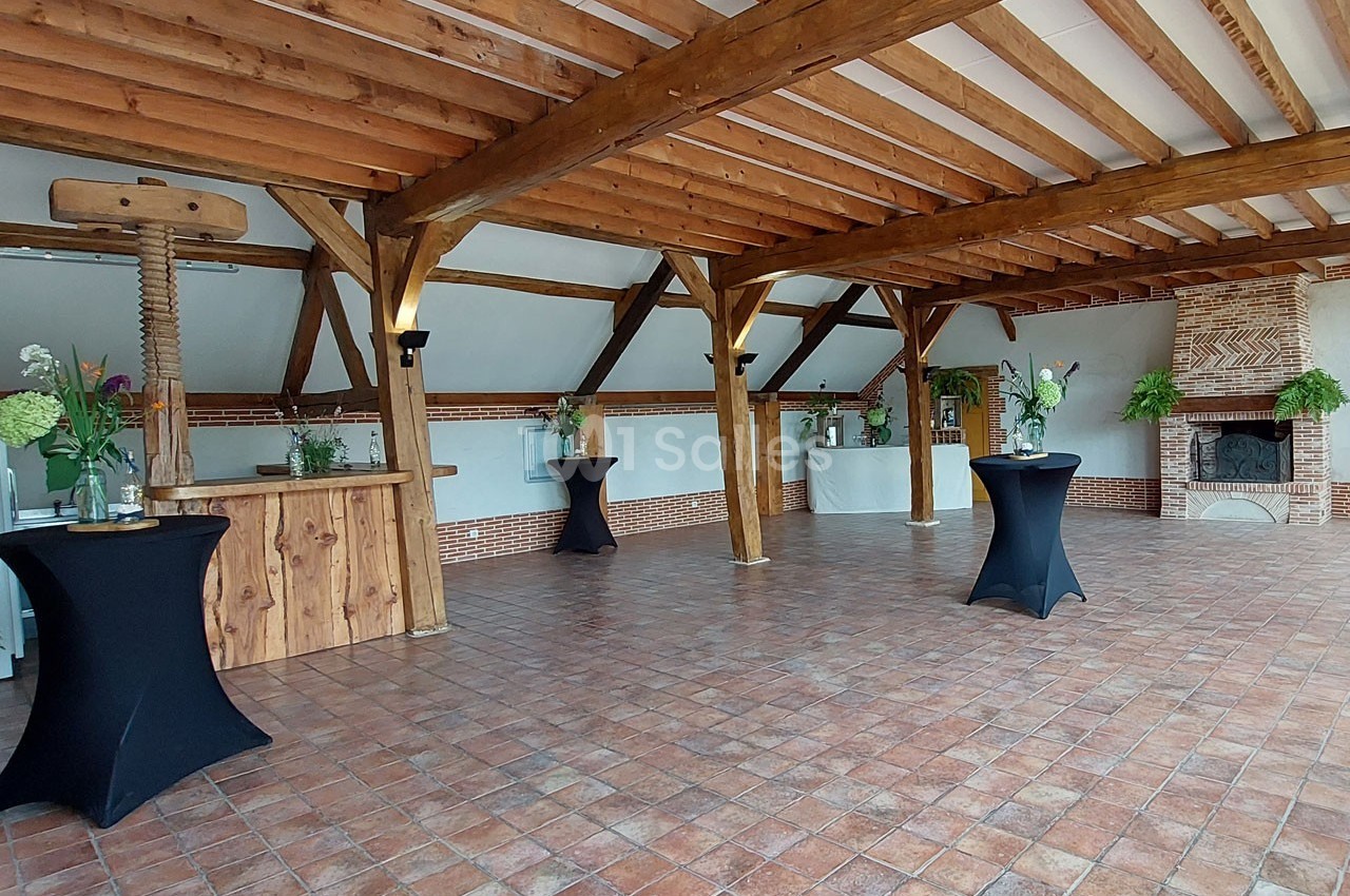 Salle avec poutres apparentes, sol en carreaux de terre cuite, tables hautes noires et cheminée en briques.