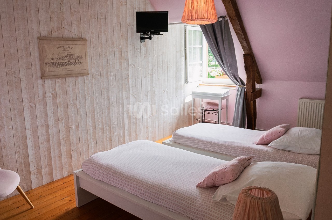 Chambre lumineuse avec deux lits simples, murs clairs, poutre apparente, fenêtre ouverte et petite télévision murale.