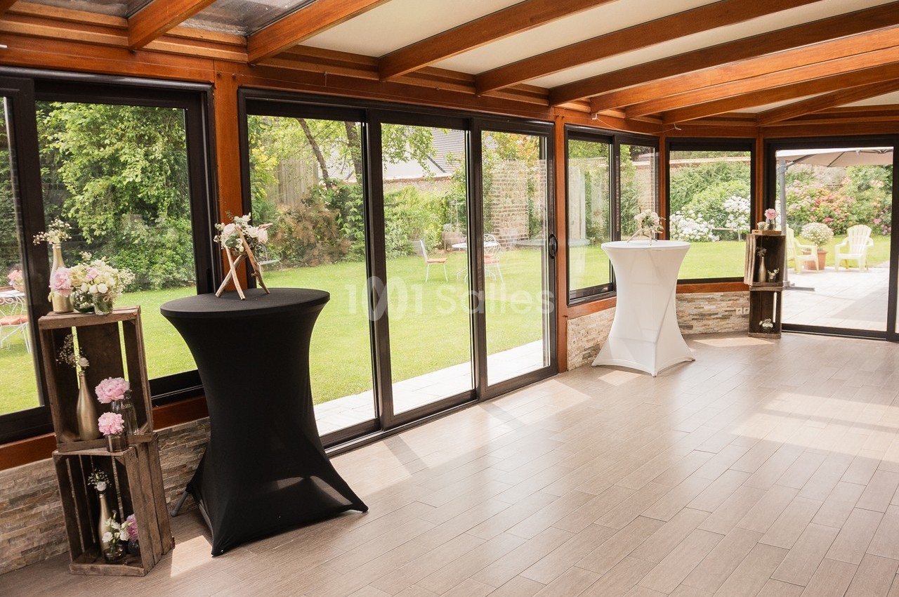 Salle lumineuse avec baies vitrées donnant sur un jardin, décorée de fleurs et de tables hautes.