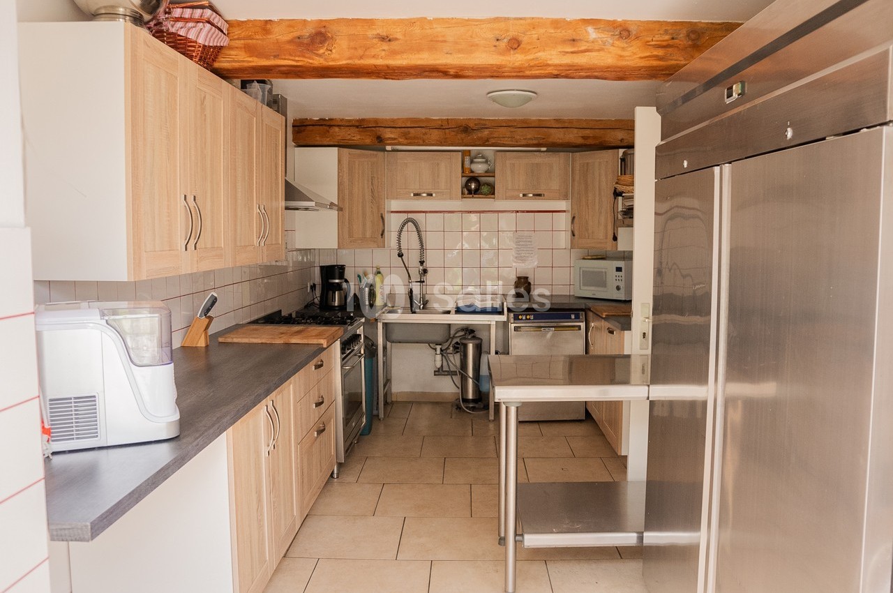 Cuisine équipée avec meubles en bois clair, plan de travail gris, électroménagers et poutres apparentes au plafond.