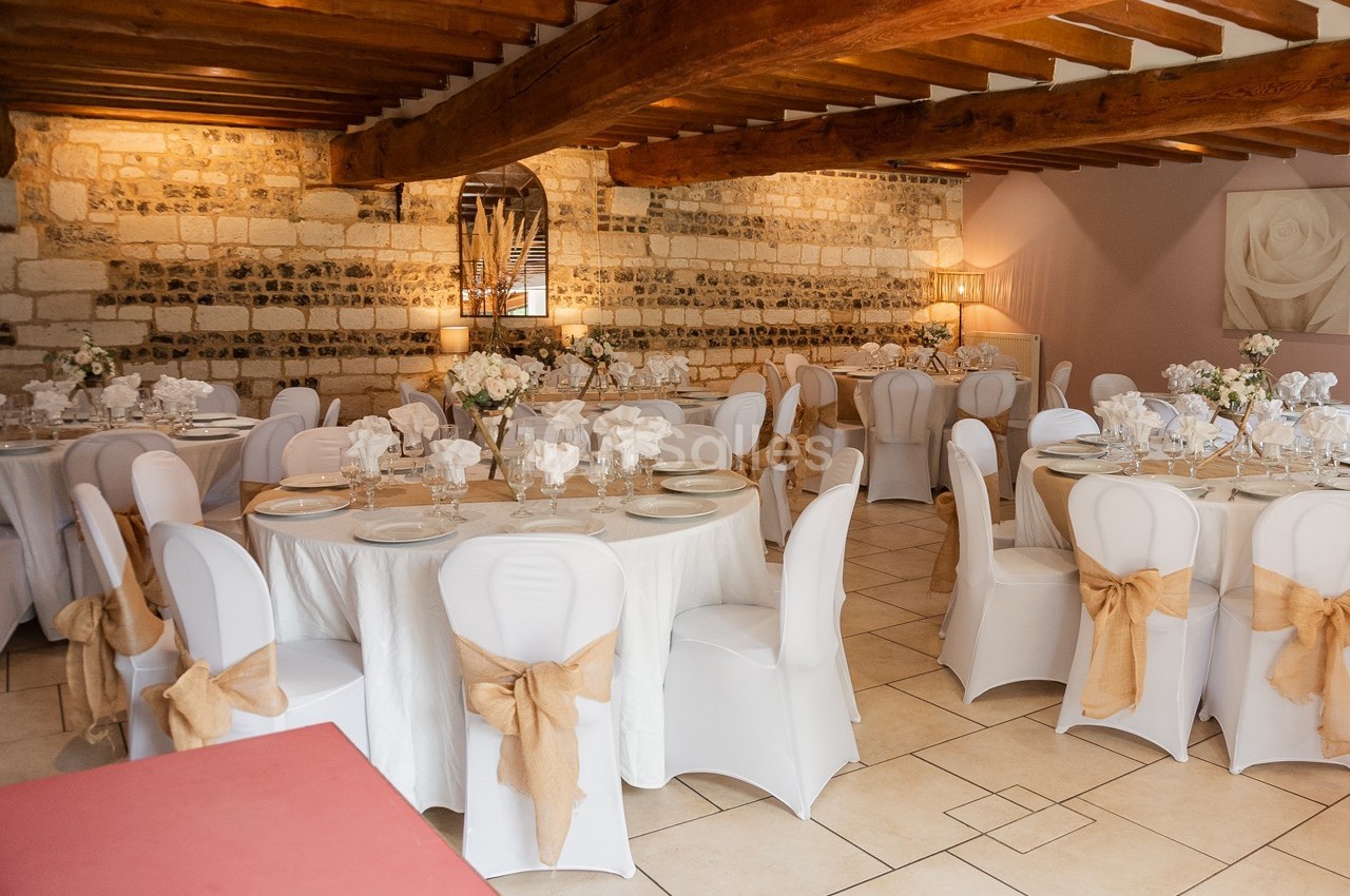 Salle de réception avec tables rondes dressées, nappes blanches, chaises décorées de nœuds beige et murs en pierre apparente.