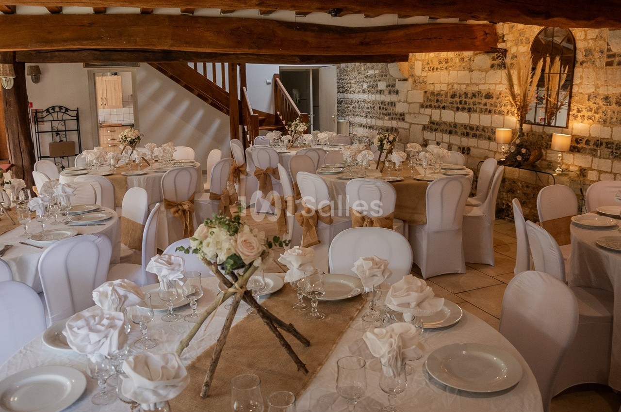 Salle de réception décorée avec des tables rondes, nappes blanches, chaises houssées et centres de table floraux.
