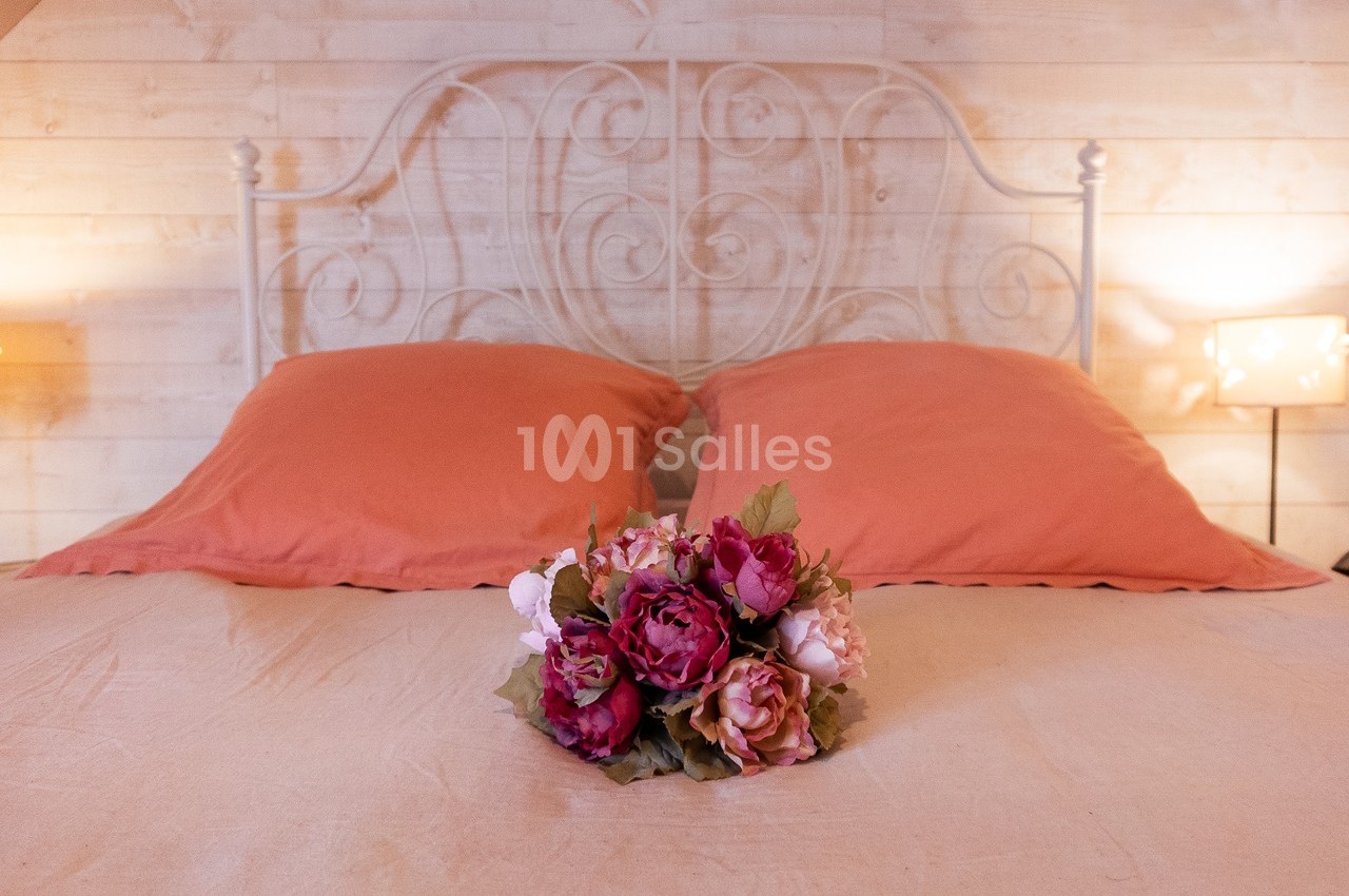 Lit avec une tête de lit en métal blanc, deux oreillers orange et un bouquet de fleurs posé sur la couverture beige.
