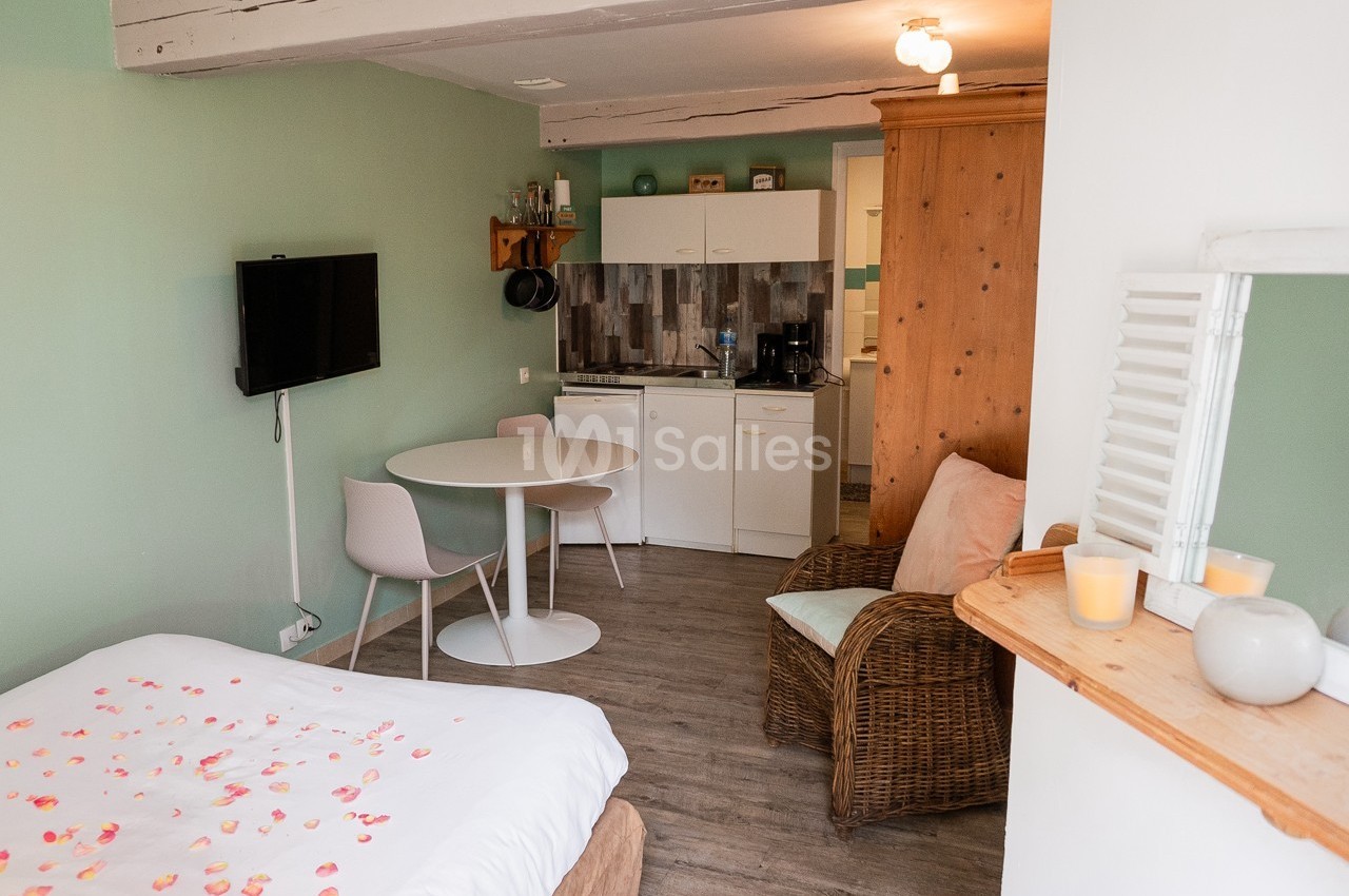 Studio lumineux avec lit, table ronde, kitchenette, fauteuil en osier et décoration simple dans des tons doux.